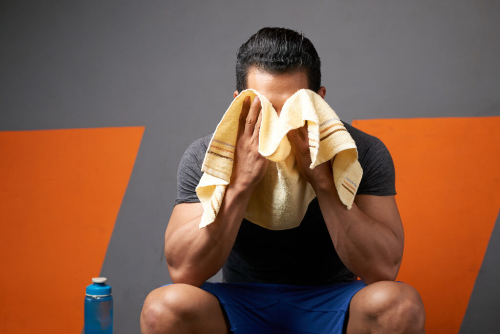 medium-shot-unrecognizable-male-athlete-wiping-sweat-with-towel-seated-gym-changing-room (1)